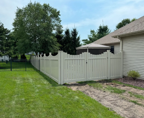 Beige vinyl fence surrounding a grassy yard with a gate, house on the right, and trees in the background.