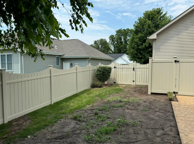 Tan vinyl fence around a yard with a gate, green grass, and beige house on a sunny day.