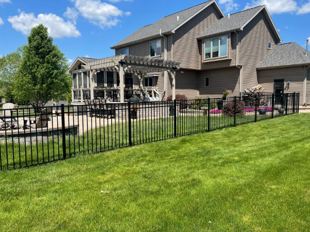 Black metal fence surrounding a green lawn in front of a multi-level house with a screened porch on a sunny day.