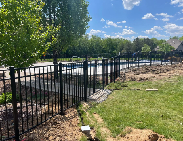 Black fence surrounding a pool area. Green grass and trees are in the background on a sunny day.