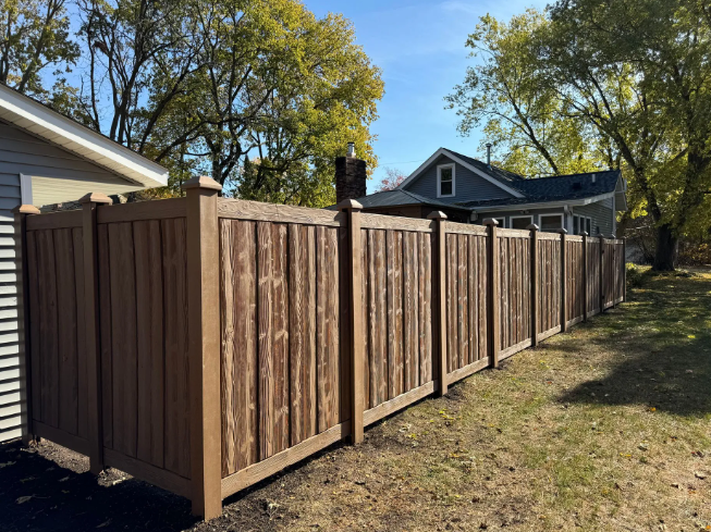 Brown privacy fence in a yard, beside a building. Green grass, trees, and blue sky.