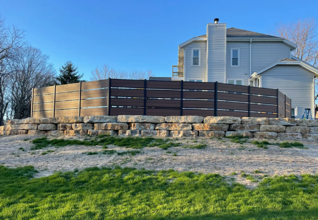 Brown privacy fence atop a stone retaining wall in front of a light blue house.
