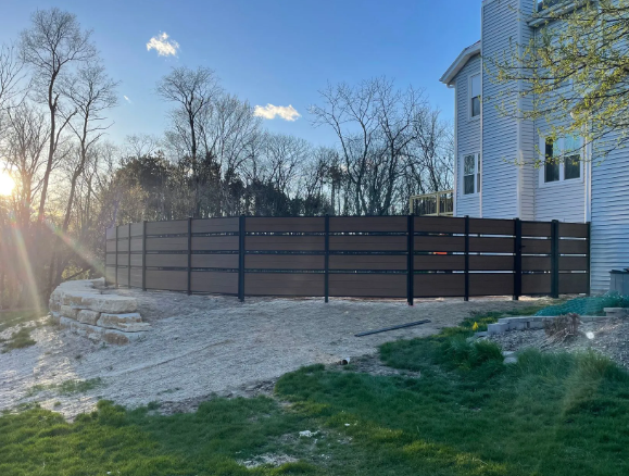 Brown and black horizontal slat fence beside a light-colored house and rocky ground, under a blue sky.