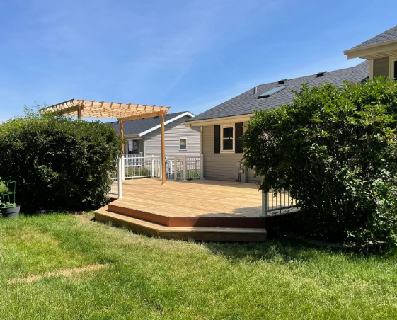 Wooden deck with pergola, surrounded by bushes and grass. House in background.