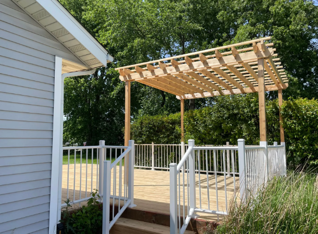 Wooden pergola on a deck with white railings next to a house with blue siding, surrounded by green trees.
