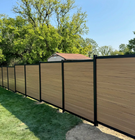 Tan horizontal privacy fence with black posts on green lawn, trees in background.