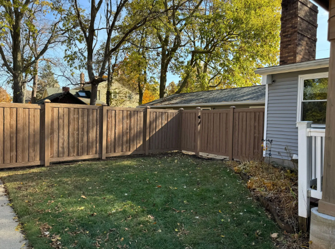 Brown fence surrounding a green lawn, next to a gray house with a chimney.