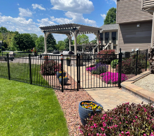 Black fence encloses a patio with flowers, landscaping, and a pergola on a sunny day.