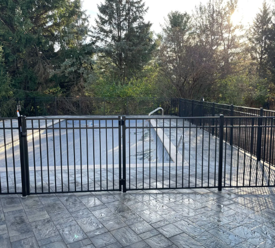 Black metal fence surrounds a swimming pool on a stone patio with trees in the background.