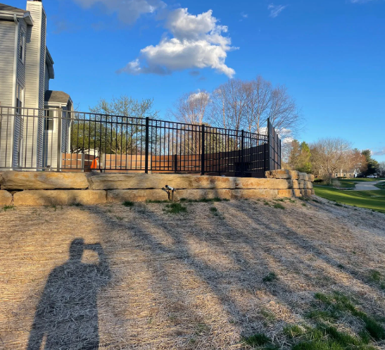 Exterior view of a house with a black metal fence on a raised patio, sunny day. Shadow of a person taking a photo.