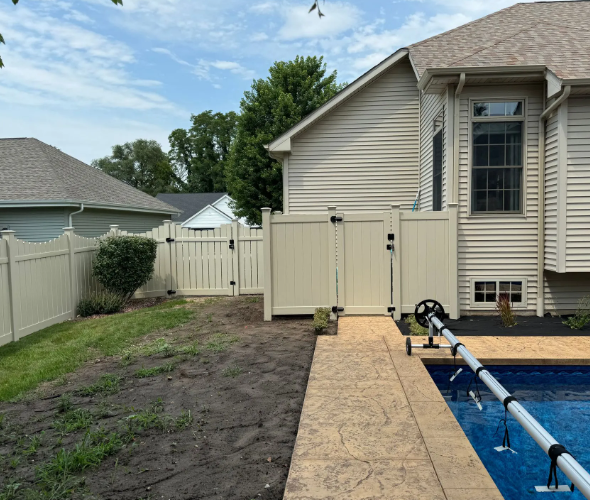 Beige fence surrounding a backyard with a pool. A gate leads to a house with beige siding and a blue sky.