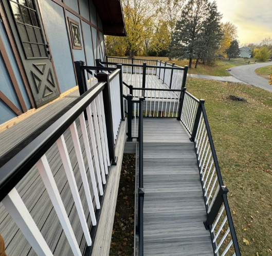 A wooden deck with stairs and railings outside a blue building, surrounded by trees and a winding road.