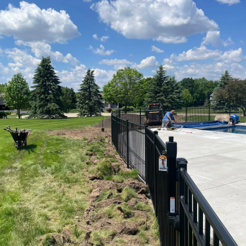 Lawn next to black fence with pool; worker by pool, blue sky, trees, wheelbarrow on lawn.