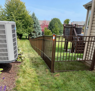 Brown metal fence bordering a backyard with an AC unit, deck, and green lawn on a sunny day.
