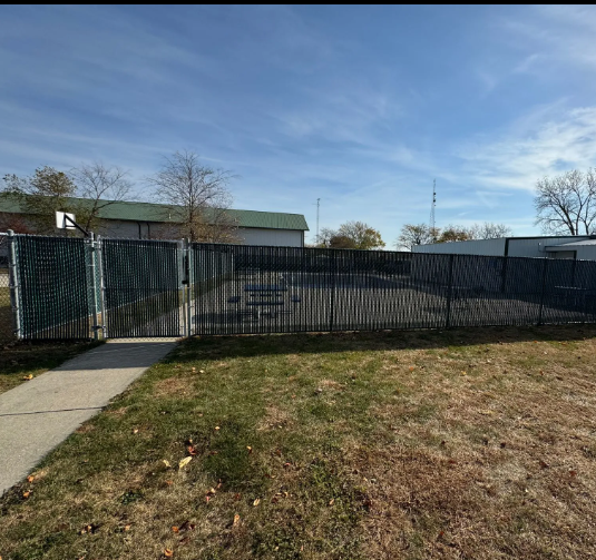 Fence encloses a grassy area with a gate. Buildings and a blue sky are in the background.