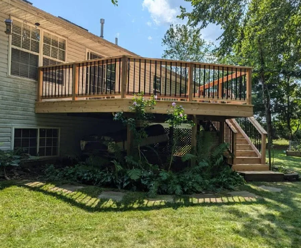 Wooden deck attached to a house with black railings and stairs surrounded by greenery on a sunny day.