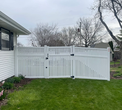 White vinyl fence with gate alongside a house on a grassy lawn.