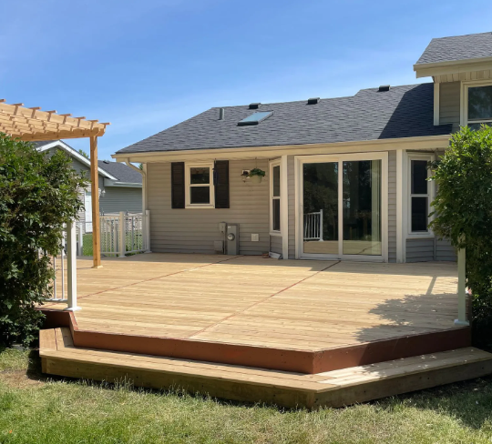 Wooden deck with steps, connected to a light-colored house with sliding glass doors and a pergola. Green lawn and blue sky.