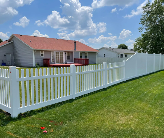 White picket fence in a grassy backyard, with houses in the background under a blue sky with clouds.