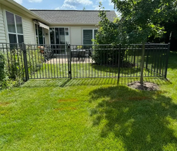 Black metal fence encloses a backyard with green grass, a small tree, and a patio with a table.