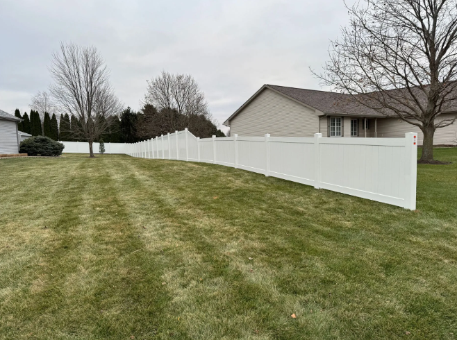 White fence lines a grassy yard with houses in the background under a cloudy sky.