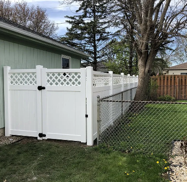 White vinyl fence with gate, bordering a grassy yard next to a building and chain-link fence.