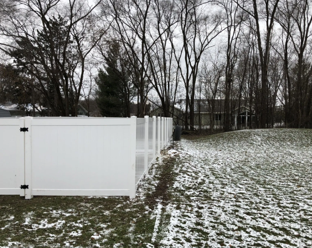 White fence bordering a snowy yard with bare trees in the background.