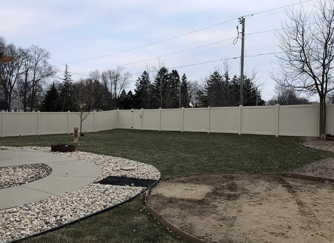 A beige privacy fence encloses a green lawn, with a concrete patio and gravel border in a backyard.