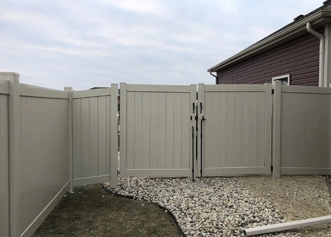 Beige vinyl fence and gate surrounding gravel area, next to a house with brown siding.