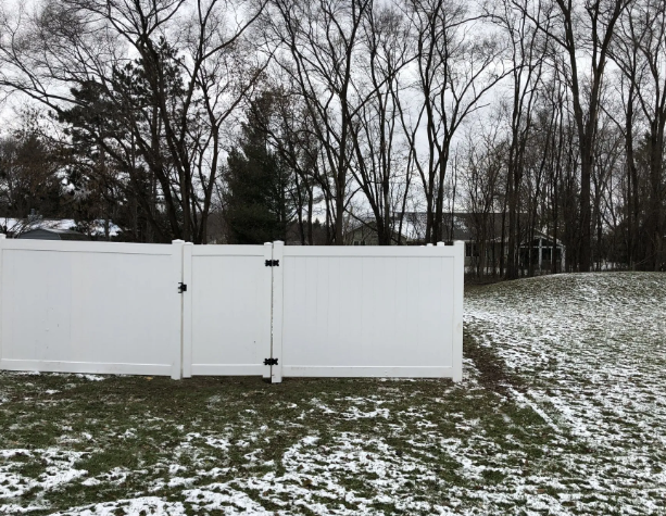 White vinyl fence with gate in a yard with patches of snow, bare trees in the background.