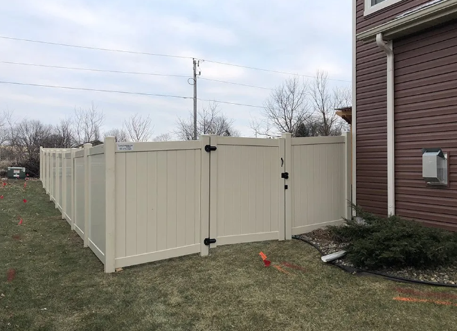 Beige vinyl fence with gate attached to a brown house with a green lawn.