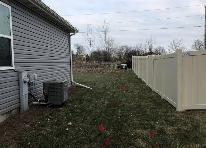 A house with gray siding has an AC unit on the side of it, next to a beige fence and a grassy yard.