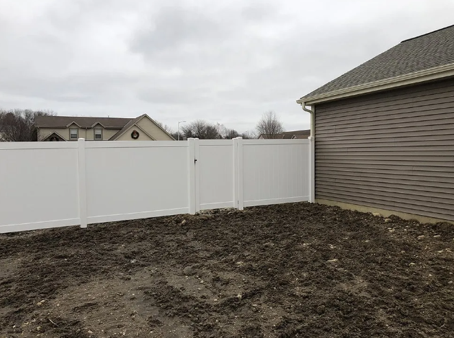 White fence enclosing a muddy yard next to a brown house with a gray roof.