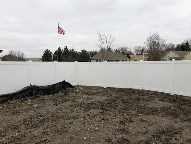 White vinyl fence surrounds a muddy yard; an American flag waves in the distance.