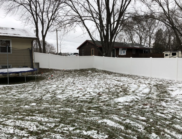 Snowy backyard with a white fence, trees, and houses in the background. A trampoline is visible.
