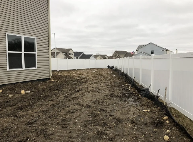Muddy backyard with a white fence, adjacent to a house. Cloudy sky.