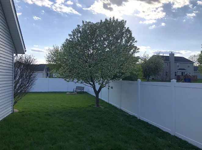 Green backyard with white fence, blooming tree, bench, and houses under a cloudy sky.