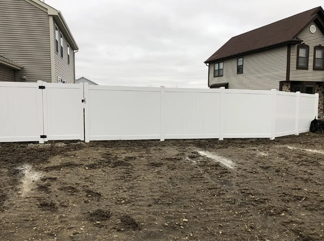 White vinyl fence in a muddy yard, between two houses under a cloudy sky.
