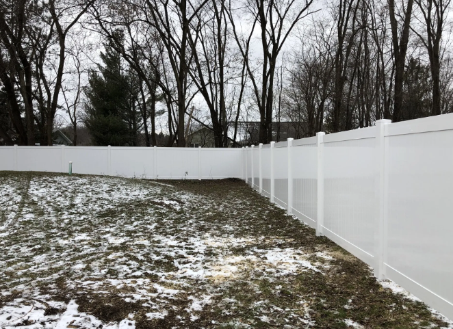 White vinyl fence surrounding a backyard with patches of snow and bare trees in the background.
