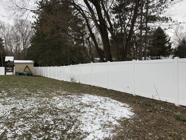 White vinyl fence bordering a backyard with patches of snow and leafless trees.