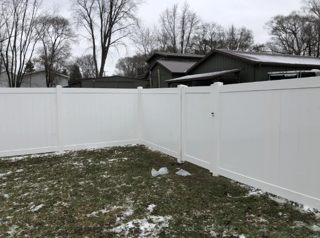 White vinyl fence enclosing a snow-covered yard, with bare trees and a dark-roofed building in the background.