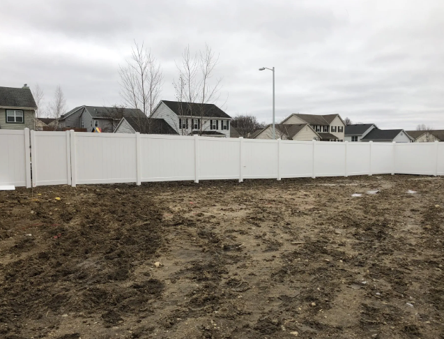 White vinyl fence surrounds a dirt lot with suburban houses in the background on a cloudy day.