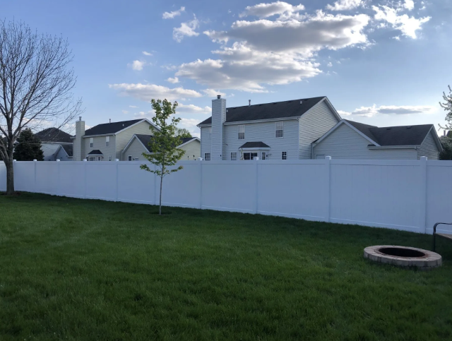 White privacy fence in backyard, with green grass and two-story houses in background under cloudy blue sky.