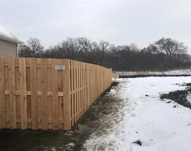 Wooden fence bordering a snowy landscape with bare trees and a cloudy sky.