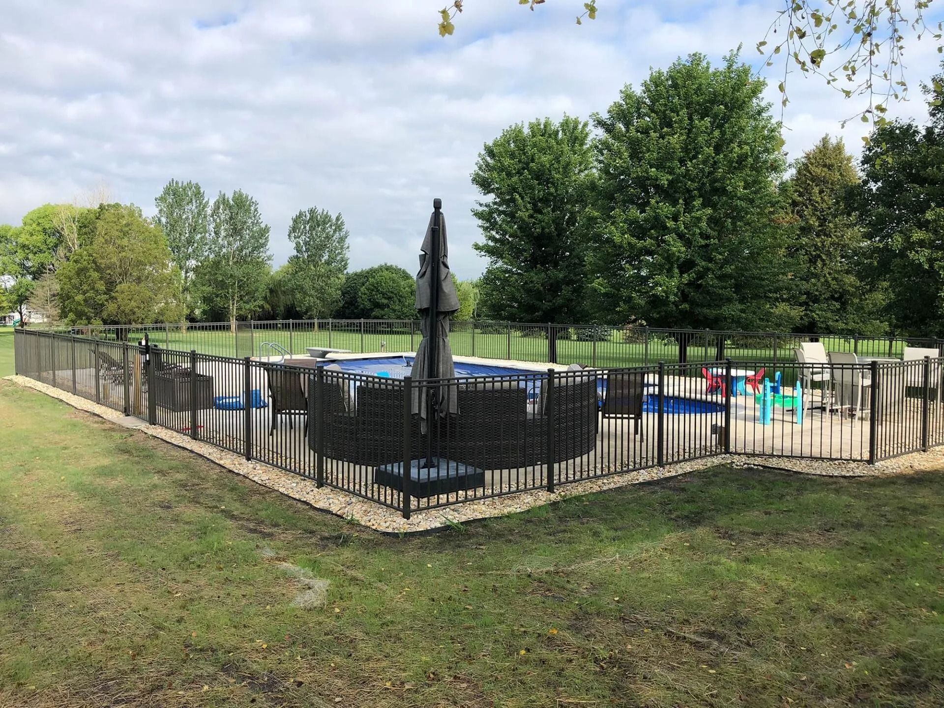 Outdoor swimming pool enclosed by black fence, with trees and cloudy sky in background.