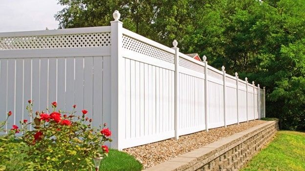 White vinyl fence with lattice top, along a retaining wall, with red roses and green trees.