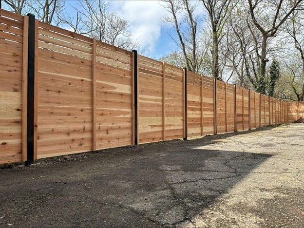 Wooden fence with black posts on a gravel surface, trees in the background, blue sky.