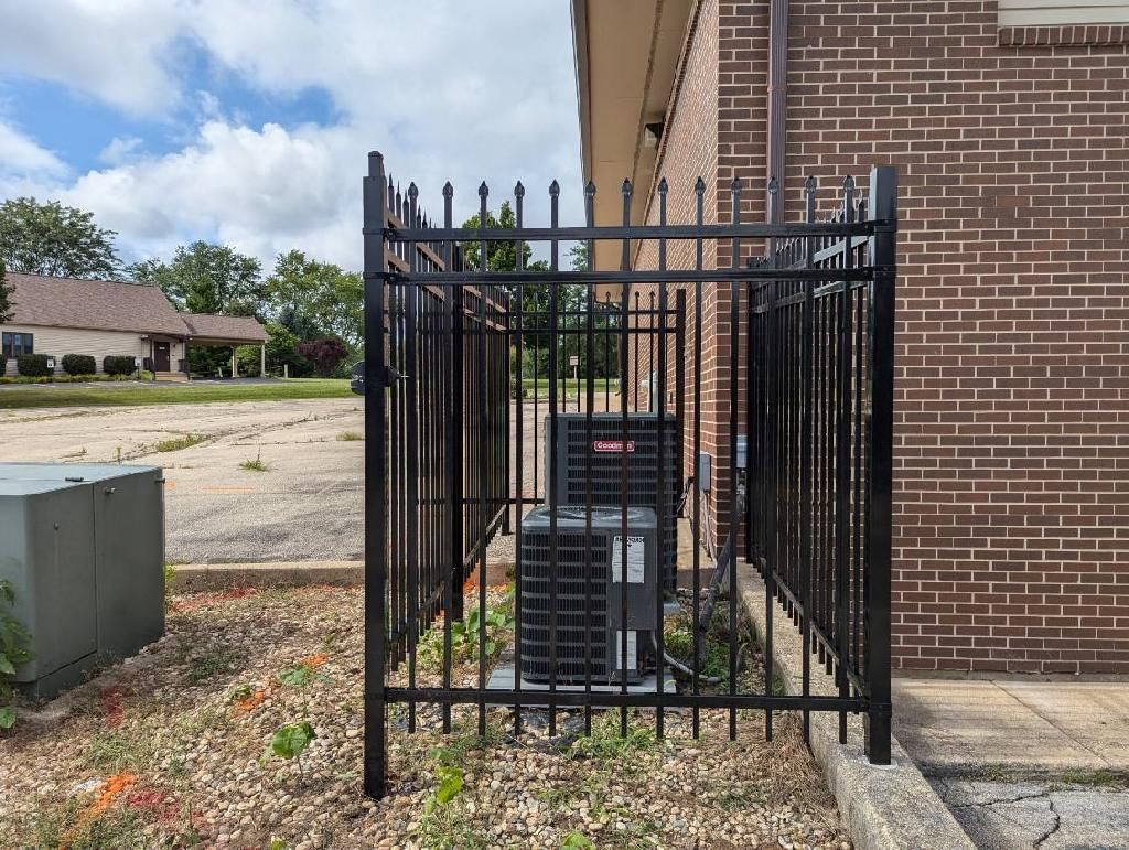 Black metal fence enclosing two air conditioning units next to a brick building.