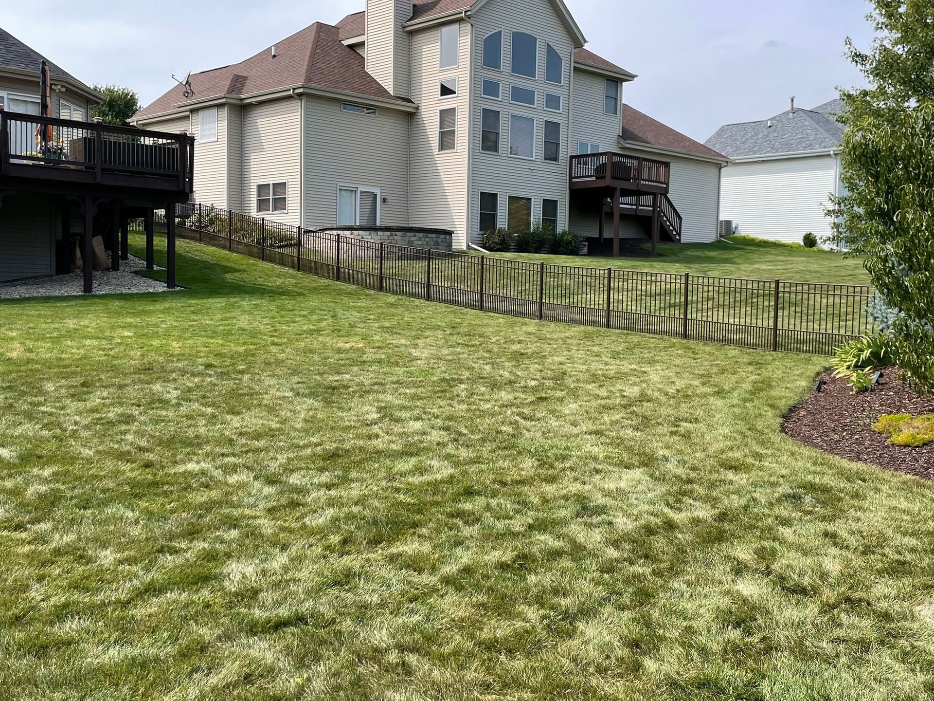 Lush green lawn slopes up to a beige house with a brown roof and a wooden fence.