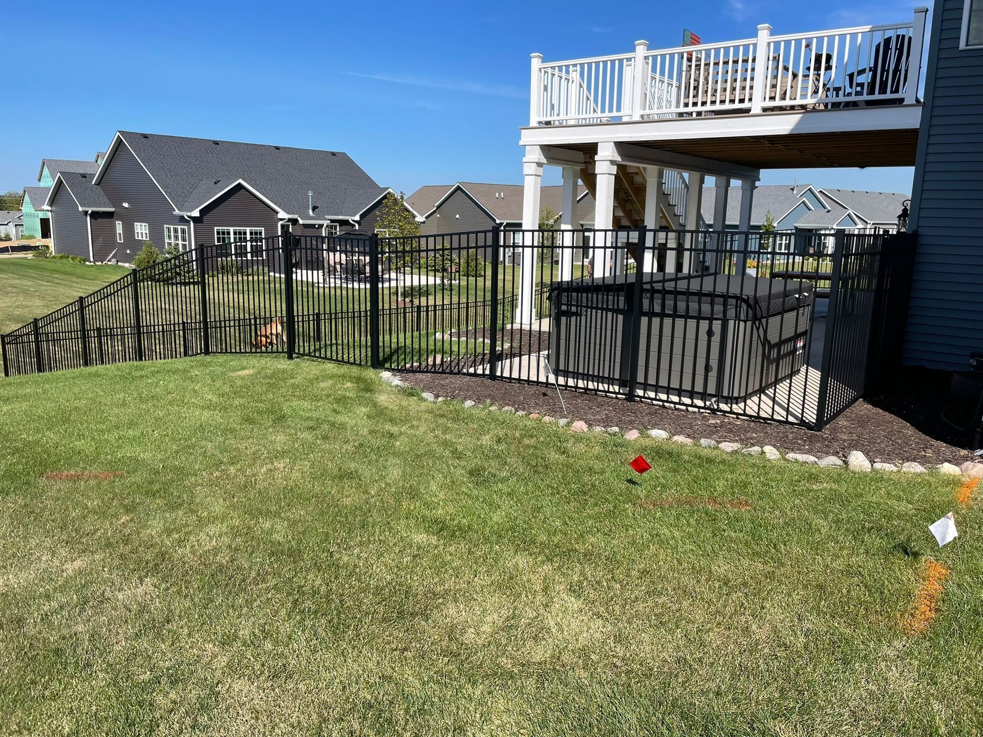 Black metal fence encloses a hot tub under a raised deck, in a grassy backyard, blue sky.